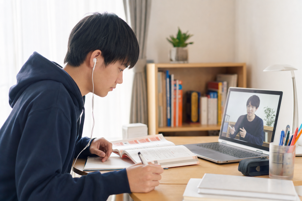 自室の机で赤本を開いて勉強する高校生と、オンライン画面越しに解説する大学生の先生